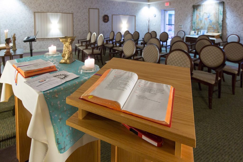 A peaceful chapel with an open Bible on the lectern, surrounded by candles and chairs arranged for a service.