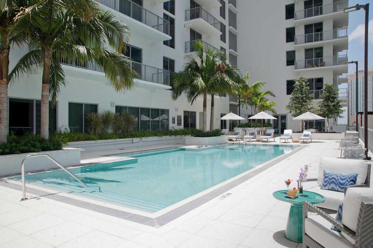 An outdoor pool area surrounded by a modern apartment building with palm trees and lounge chairs.