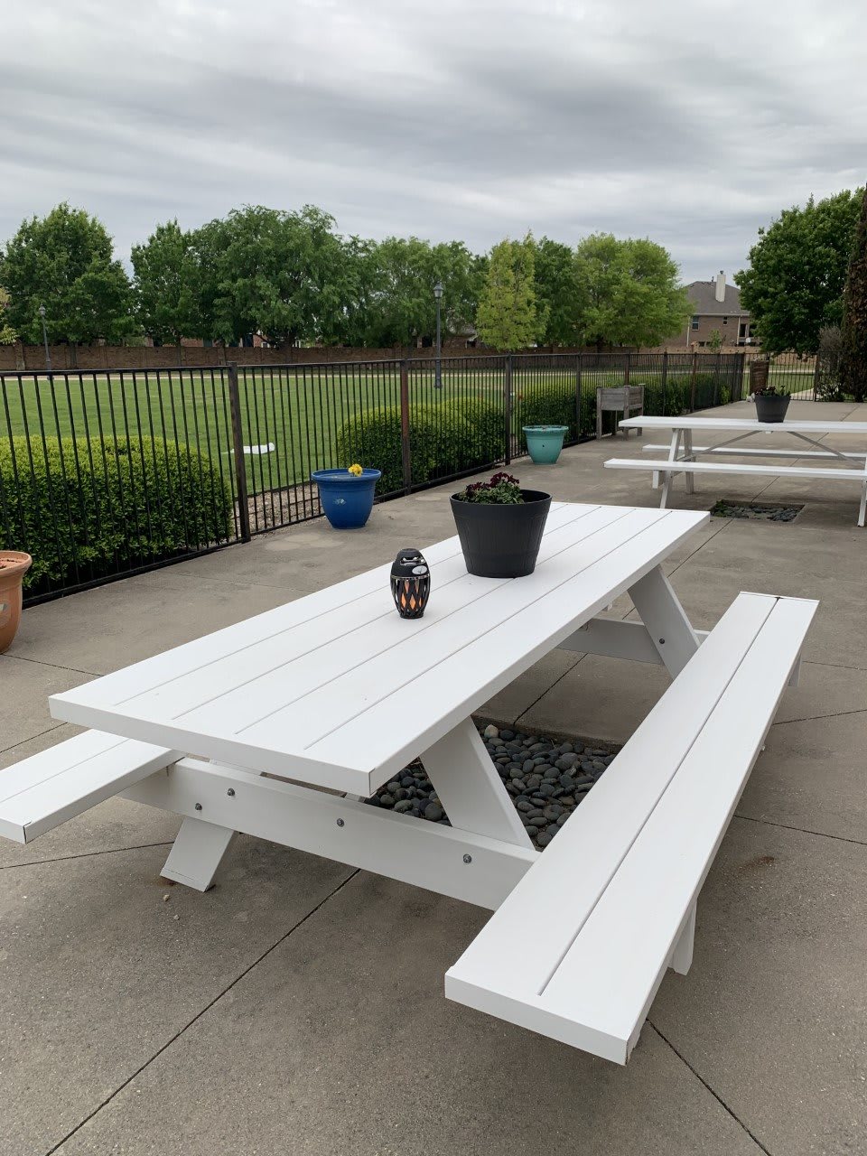 A white picnic table and benches on a patio, adorned with potted plants, overlooking a green field.