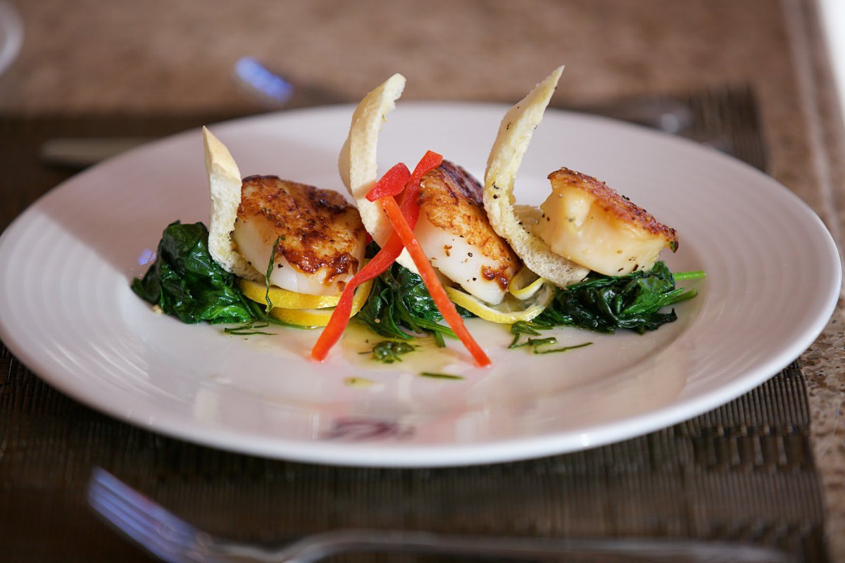 A gourmet dish featuring seared scallops atop a bed of spinach and garnished with red pepper and decorative crisps.