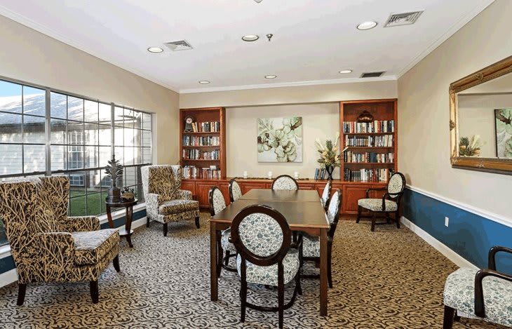 A cozy library room with bookshelves, a table, and chairs arranged on a carpeted floor.