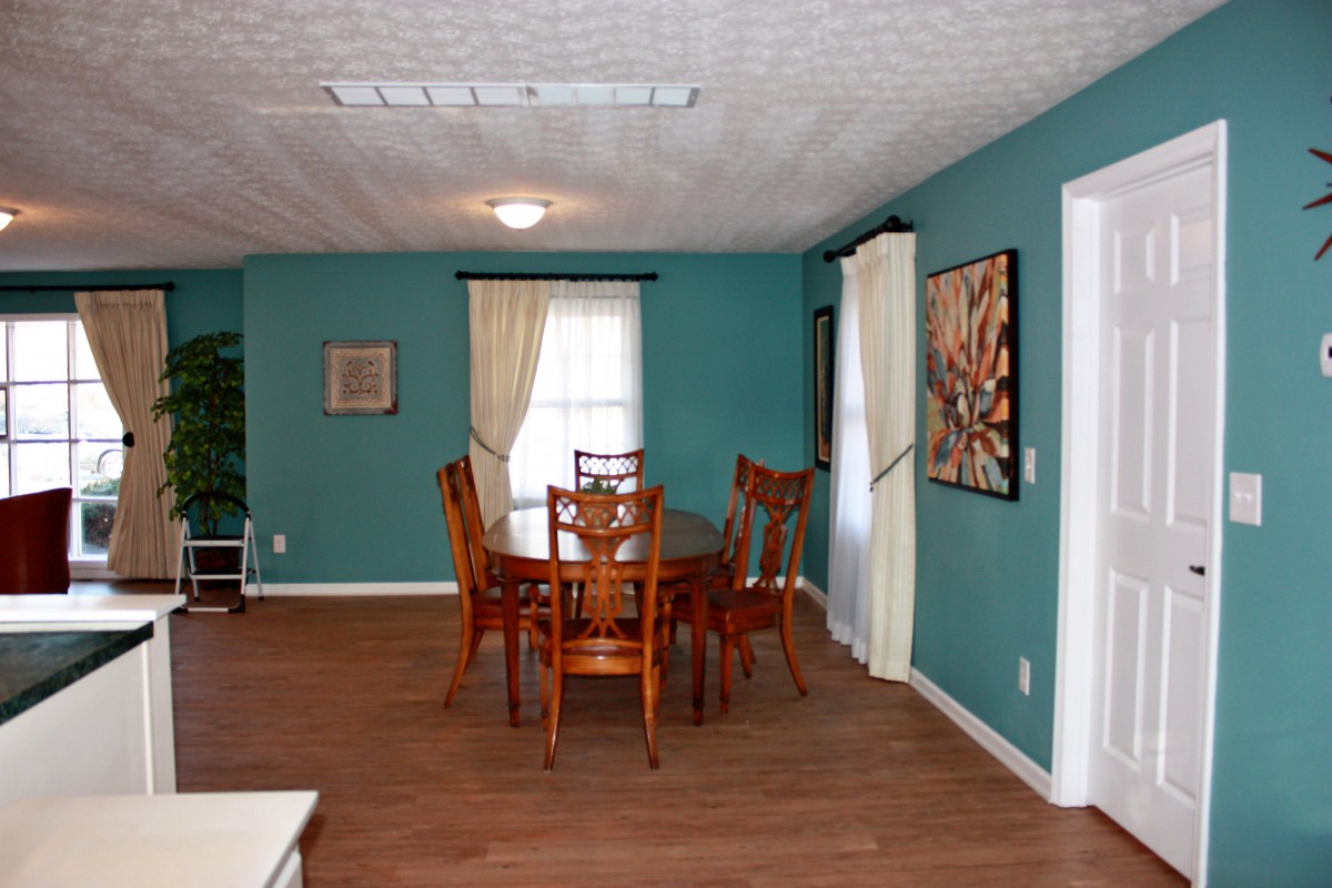 A cozy dining room with teal walls features a wooden table surrounded by chairs, hardwood flooring, and adorned with artwork and curtains.
