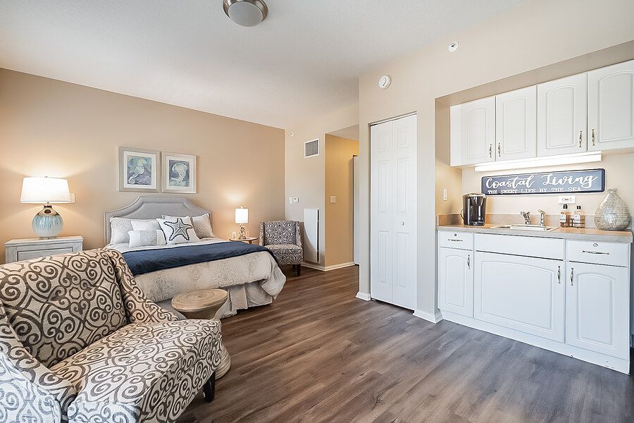 A cozy bedroom with a bed, chair, and kitchenette area, featuring wood flooring and wall artwork.