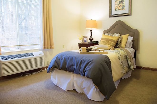 A cozy bedroom with a neatly made bed, warm lighting from a lamp, and a large window letting in natural light.