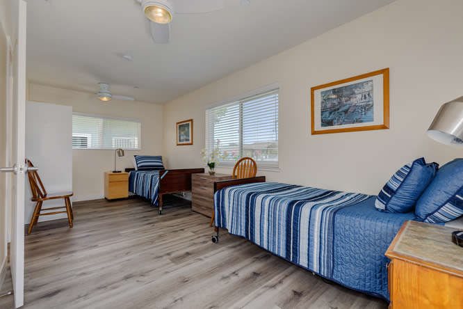 A bright and airy bedroom with a single bed featuring blue striped bedding, a desk and chair, large windows, and artwork on the walls.