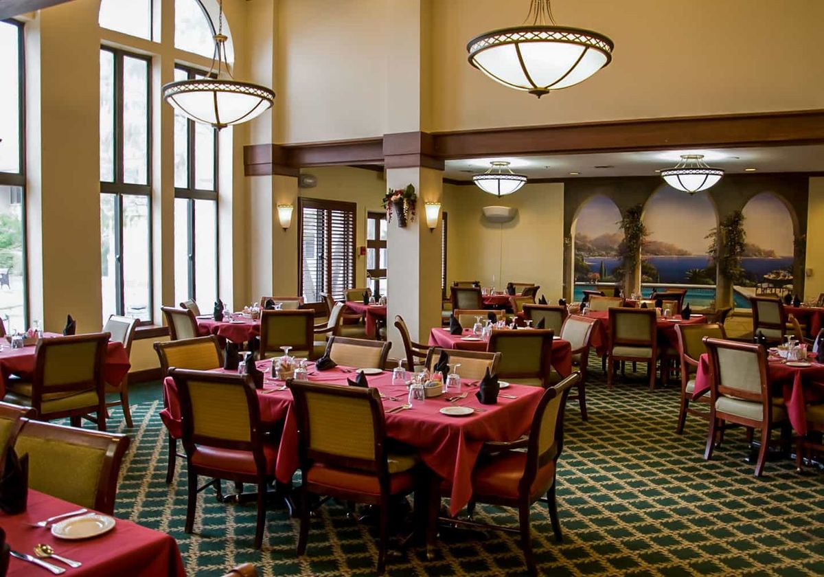 An elegant dining area with multiple tables and chairs, illuminated by large windows and chandeliers.