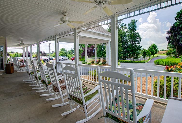 A row of white rocking chairs on a covered porch overlooking a suburban street.