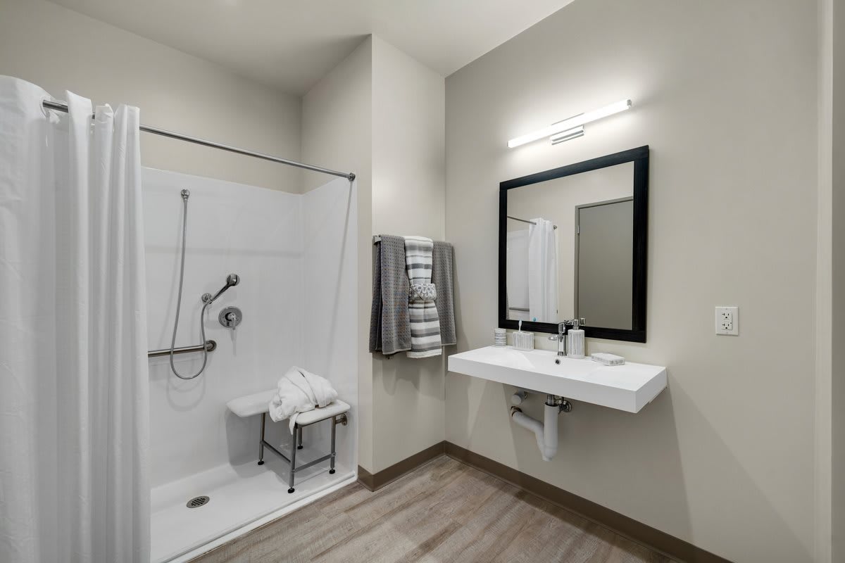 A modern, accessible bathroom featuring a walk-in shower with a shower chair, a large mirror above a sink, and neatly arranged towels.