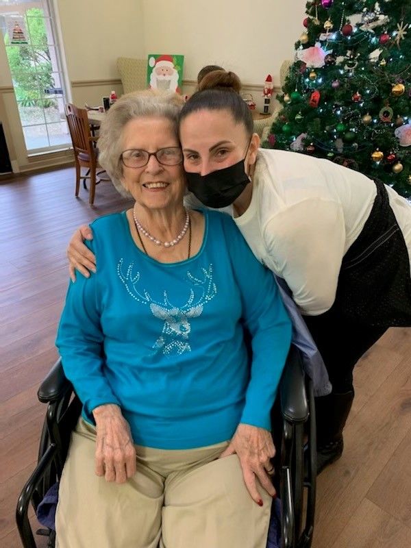 An elderly woman sitting in a wheelchair is hugged by a younger woman wearing a mask, with a Christmas tree in the background.