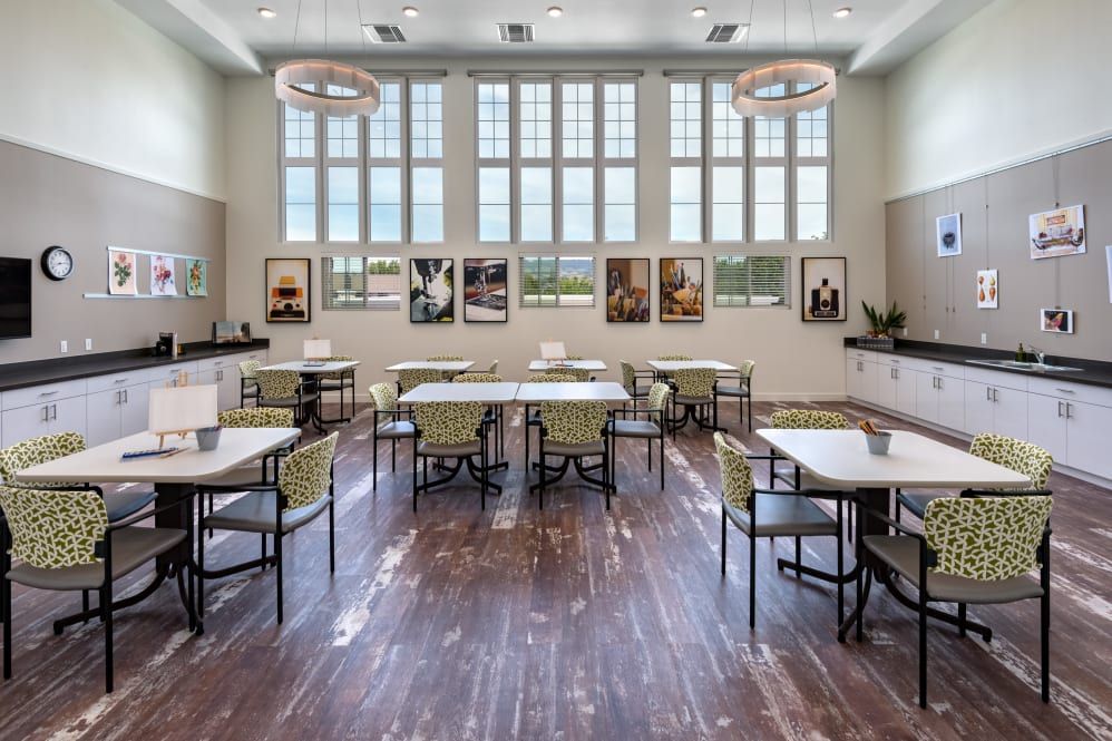 A modern dining room with large windows, tables, and chairs arranged neatly, featuring artwork on the walls and wooden flooring.