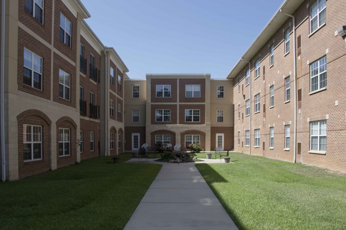 A three-story apartment building with a brick facade, featuring a central pathway and surrounding lawn.