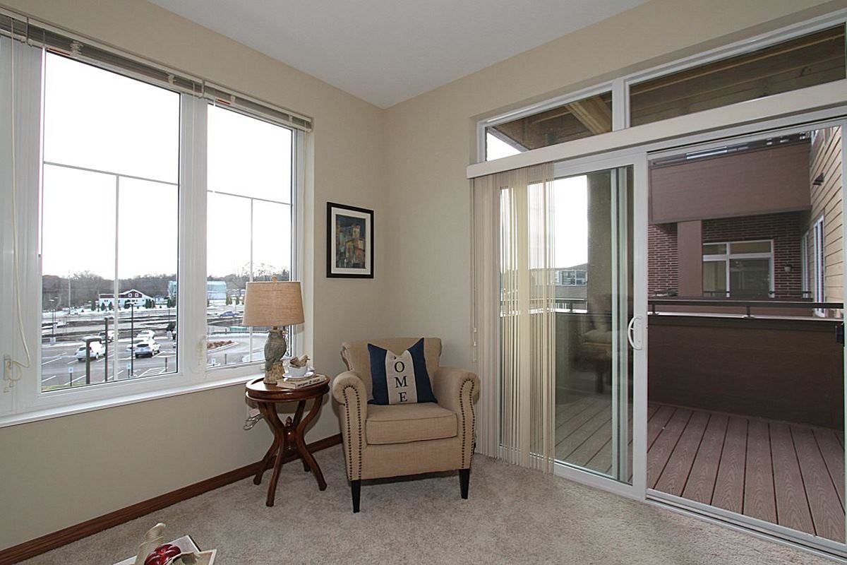 A cozy living room corner featuring a comfortable chair next to a small side table with a lamp, large windows, and a sliding glass door leading to a balcony.