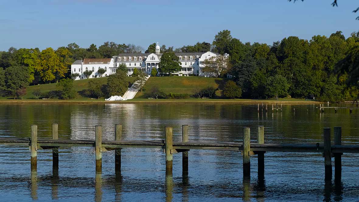 A grand mansion overlooking a serene lake with a wooden pier in the foreground.