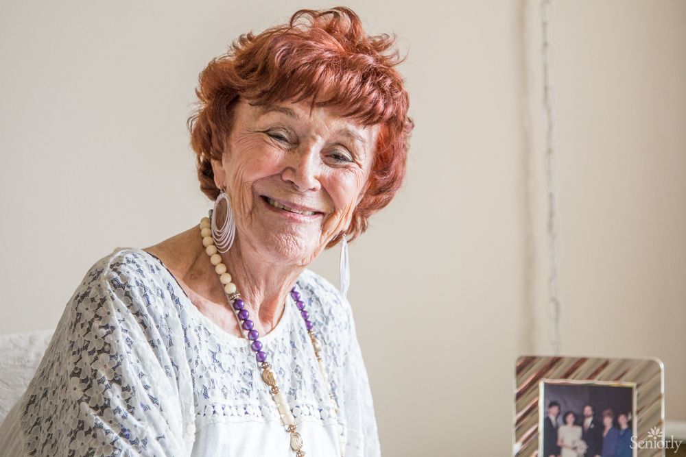 A smiling elderly woman with red hair sitting indoors.