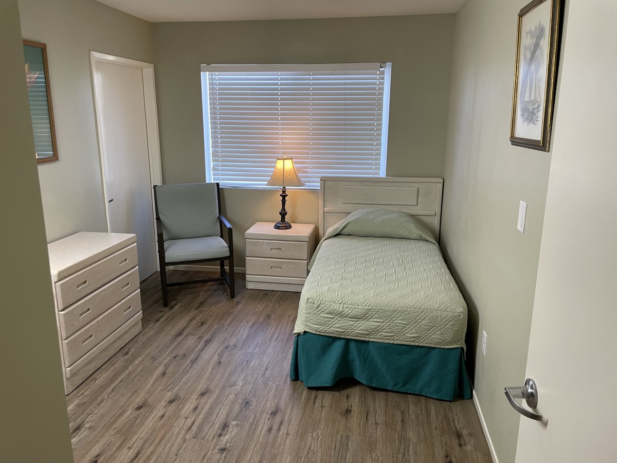 A cozy bedroom featuring a single bed, nightstand with a lamp, a chair, and a dresser on a wooden floor.