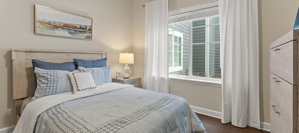 A cozy bedroom with a neatly made bed, a bedside lamp, and curtains framing a large window.