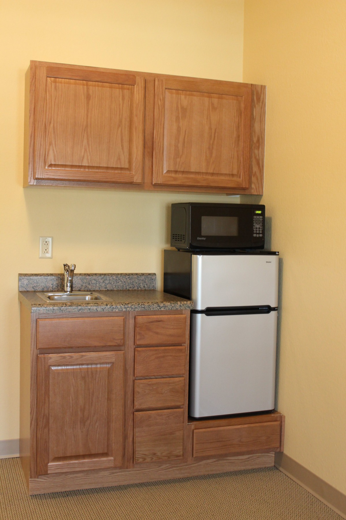 A compact kitchenette featuring wooden cabinets, a microwave on top of a mini fridge, and a small sink with a countertop.