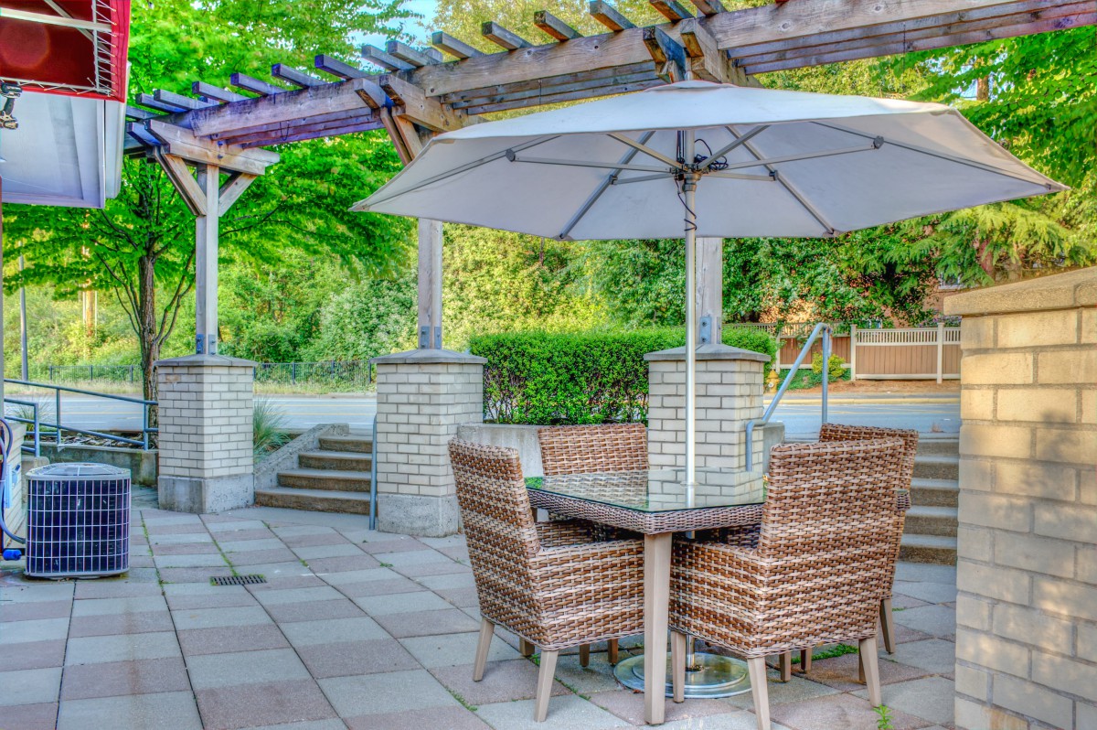 A cozy patio setup with wicker chairs and a table under a pergola, shaded by an umbrella in a garden setting.