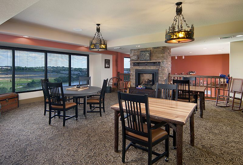 A cozy dining room with rustic decor, featuring wooden tables and chairs, a stone fireplace, chandeliers, and large windows overlooking a scenic view.