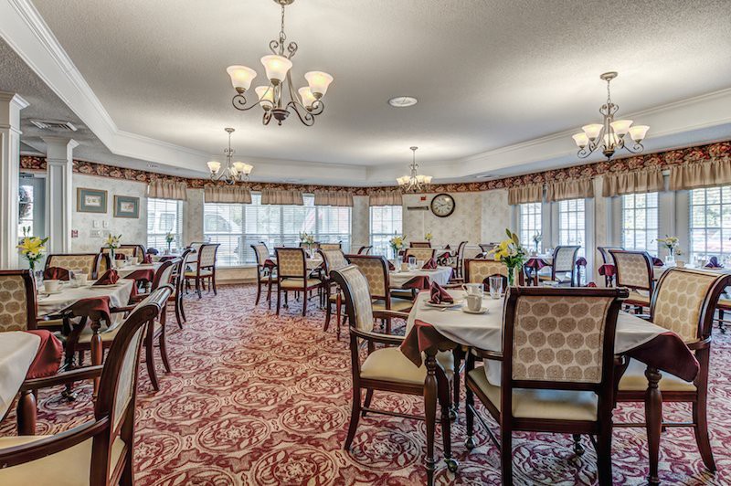 An elegant dining room with multiple tables and chairs, illuminated by chandeliers, featuring carpeted flooring and large windows.