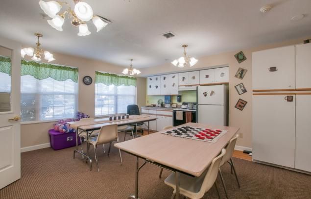 A cozy classroom setup with tables and chairs, featuring a kitchenette area and natural lighting from the windows.
