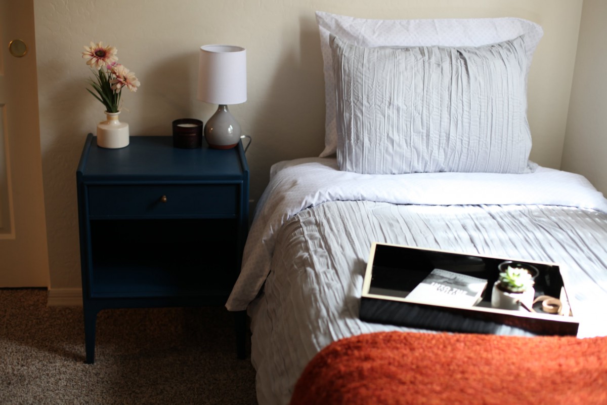 A cozy bedroom with a neatly made bed and a nightstand featuring a lamp and flowers.
