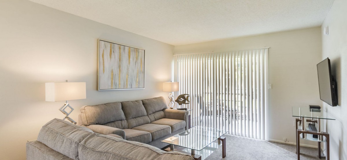 A modern living room with a comfortable grey sofa and contemporary artwork, softly illuminated by a table lamp near window blinds.