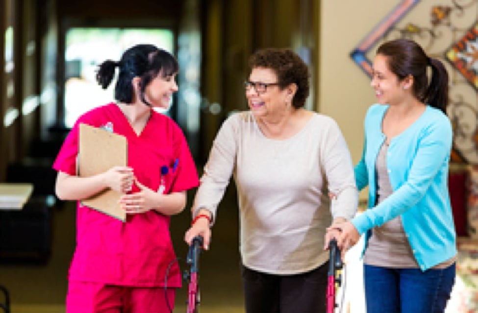 A healthcare professional assists an elderly woman using a walker, accompanied by another caregiver.