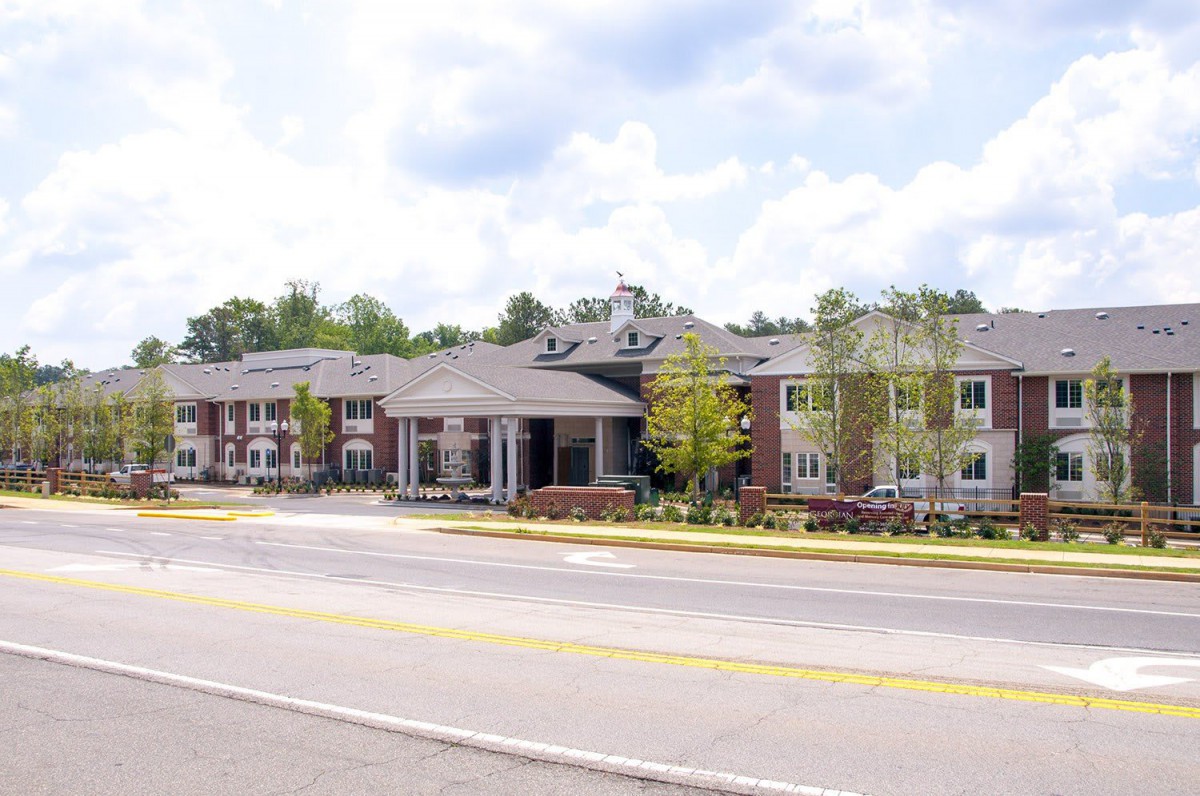 A large senior living facility with a landscaped entrance is seen from across the road on a cloudy day.