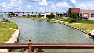 A scenic view of a park with a lake, bridge, and buildings under a partly cloudy sky.