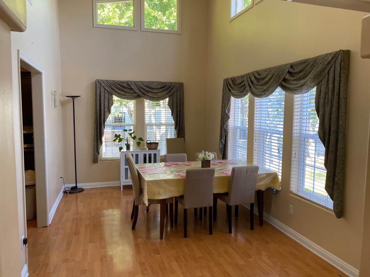 A bright dining room with a table set for six, featuring large windows with decorative curtains and wood flooring.