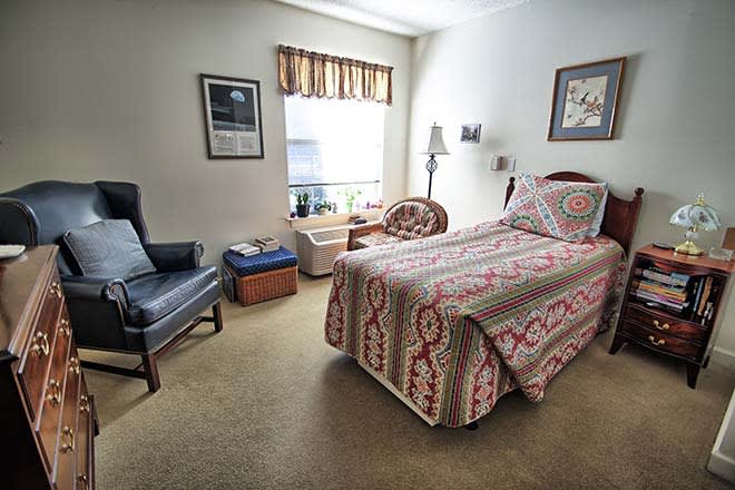 A cozy bedroom with a single bed featuring a decorative quilt, an armchair, and natural light from the window.