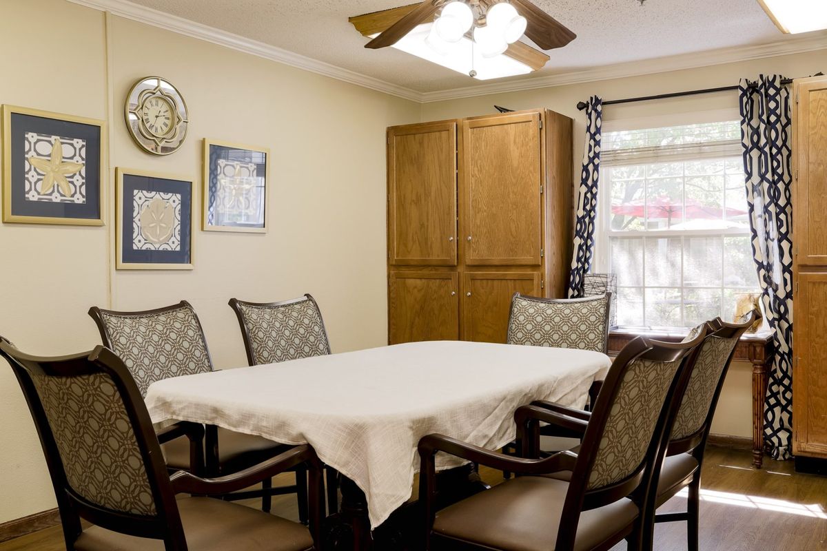 A cozy dining room with a table covered by a white cloth and surrounded by wooden chairs, featuring wall artwork and a ceiling fan overhead.