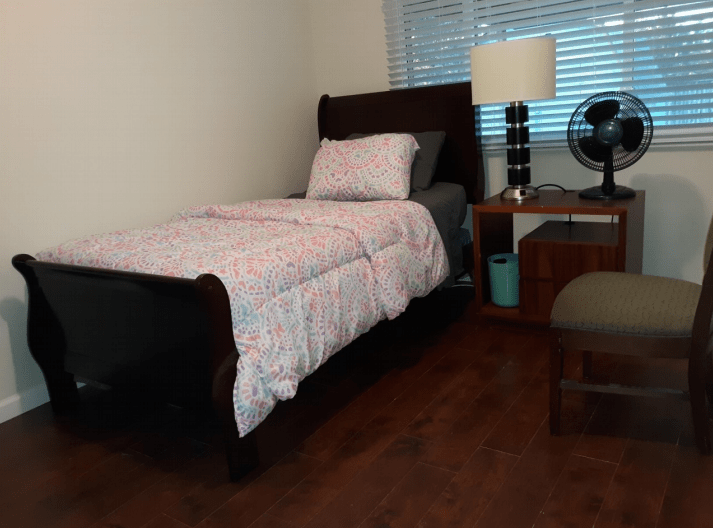 A neatly arranged bedroom with a single bed, nightstand, lamp, and fan.
