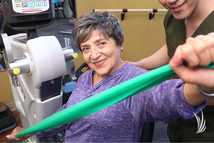 An elderly woman smiling while exercising with a resistance band, assisted by a therapist.