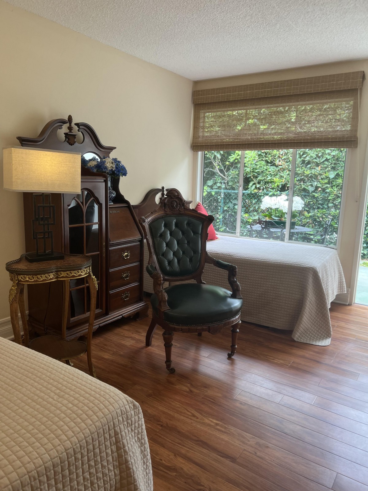 A cozy bedroom featuring a large armoire, an elegant green chair, a bedside lamp, and a window with garden view.