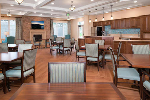 A modern dining area with wooden floors, featuring tables and chairs, adjacent to a kitchen space.