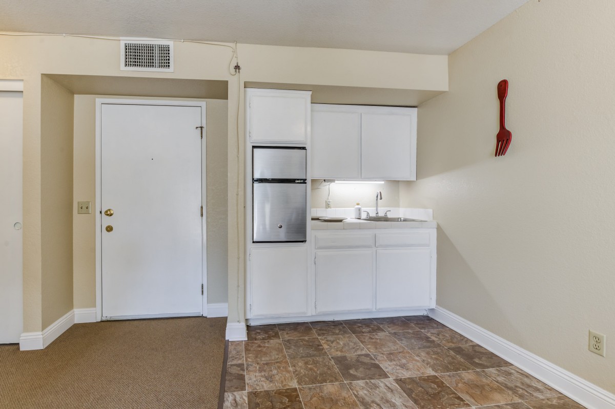 A small apartment interior featuring a kitchenette with white cabinets and a compact fridge, adjacent to the main entrance door.