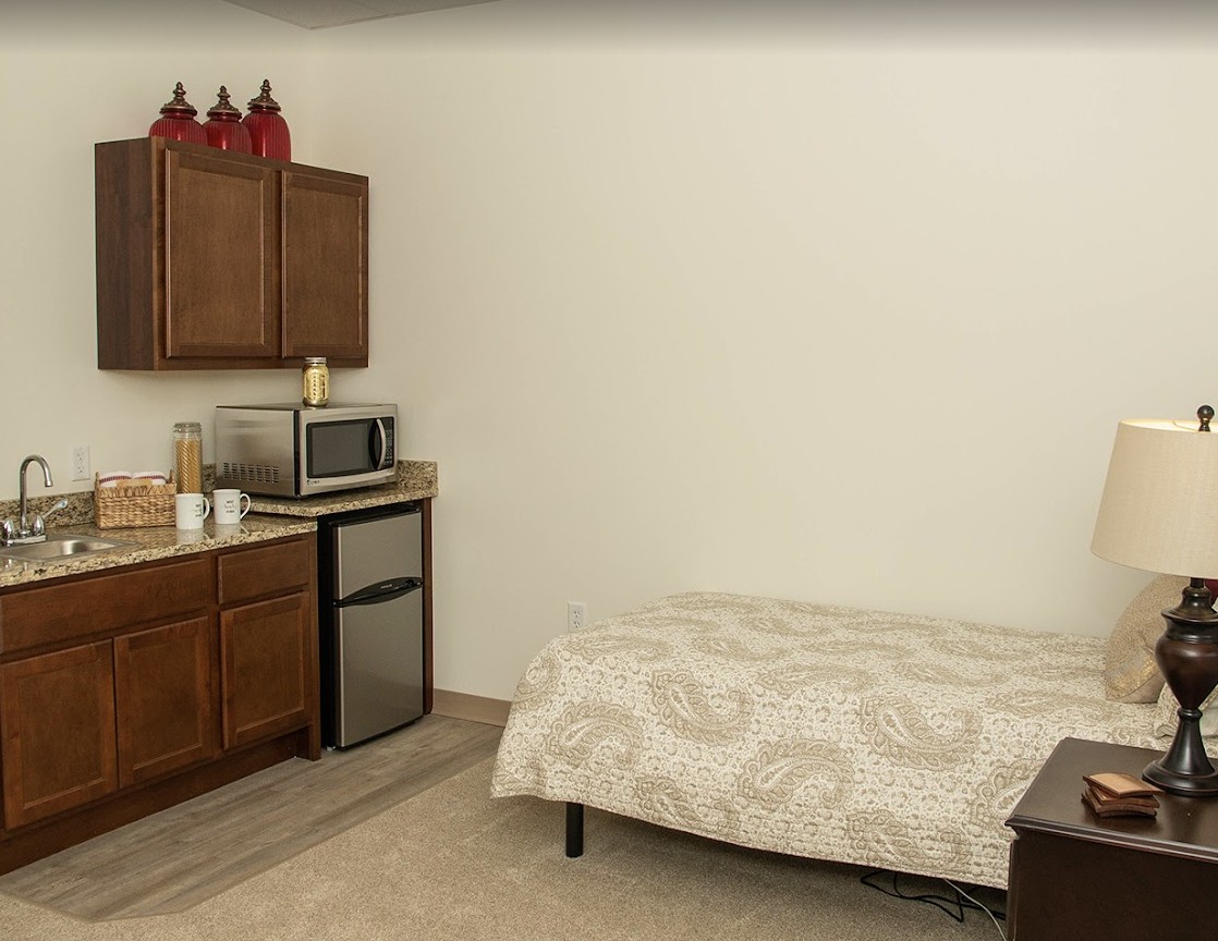 A small apartment room featuring a bed and a kitchenette with cabinets, a microwave, and a mini fridge.