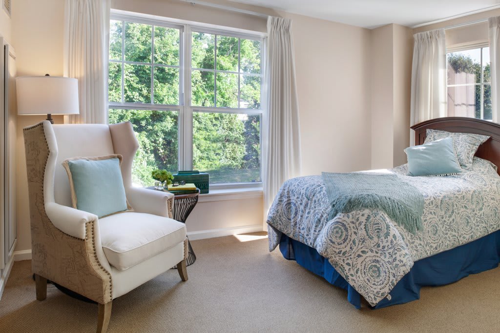 A cozy bedroom with natural light, featuring a plush armchair and a neatly made bed near a large window.