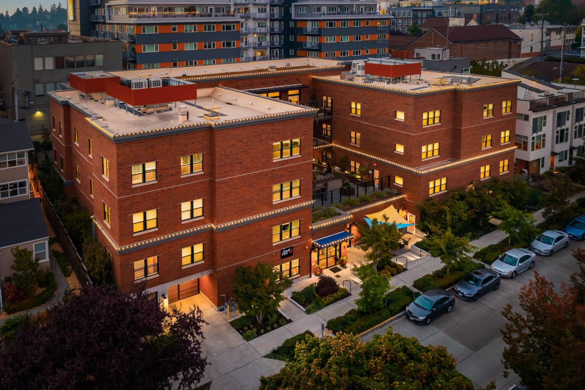 Aerial view of a brick building at dusk with lights on, surrounded by trees and parked cars.