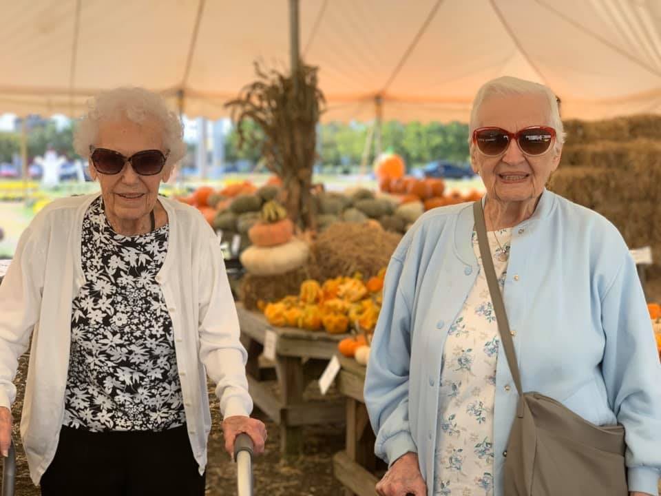 Two elderly women wearing sunglasses at an outdoor market with pumpkins.