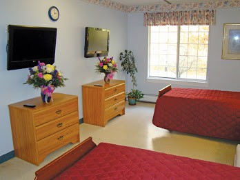 A hospital room with two beds, a television, dressers with flowers, and a window.