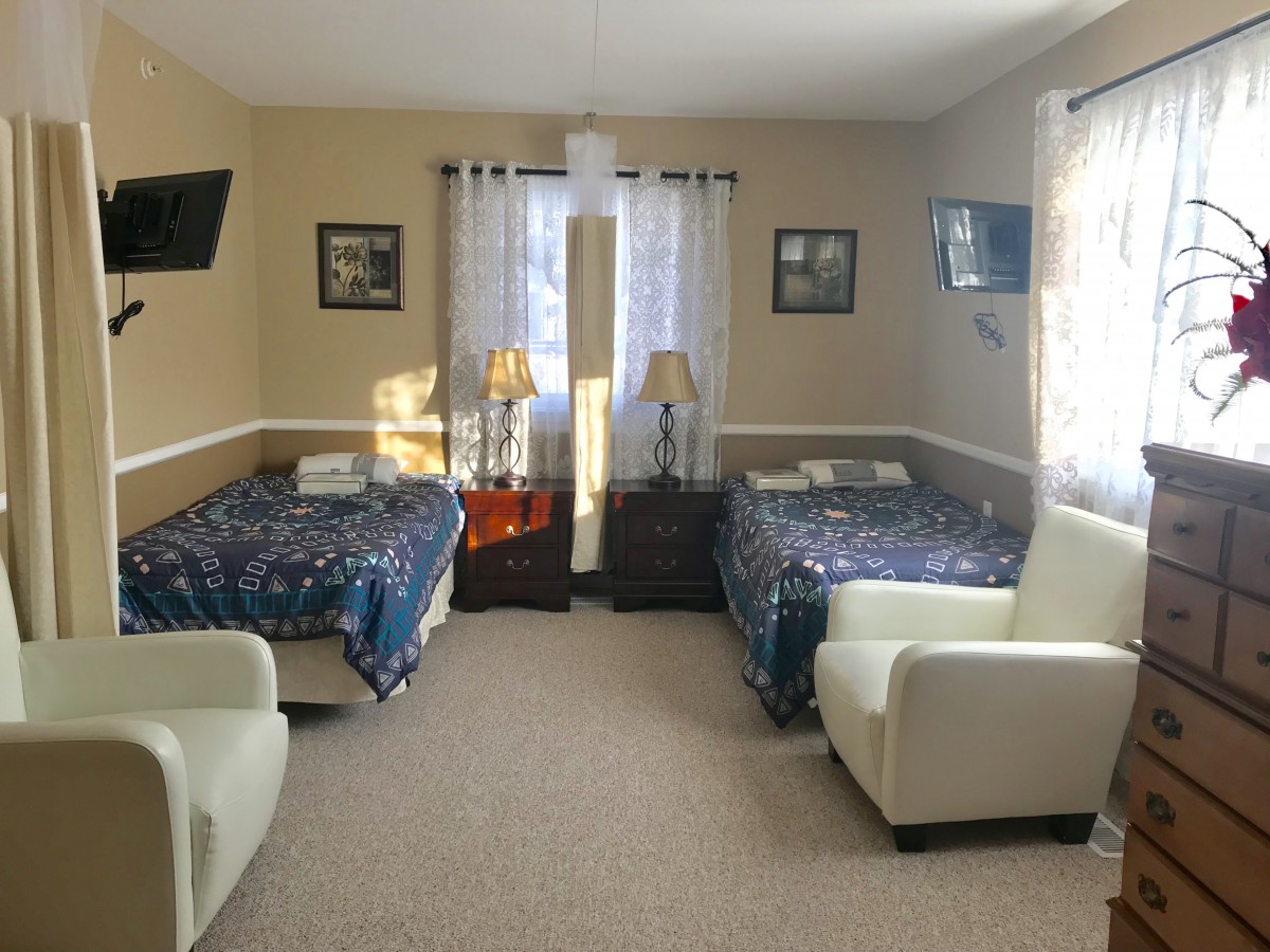 A cozy bedroom with two beds, each accompanied by a chair and television, illuminated by natural light through the window.