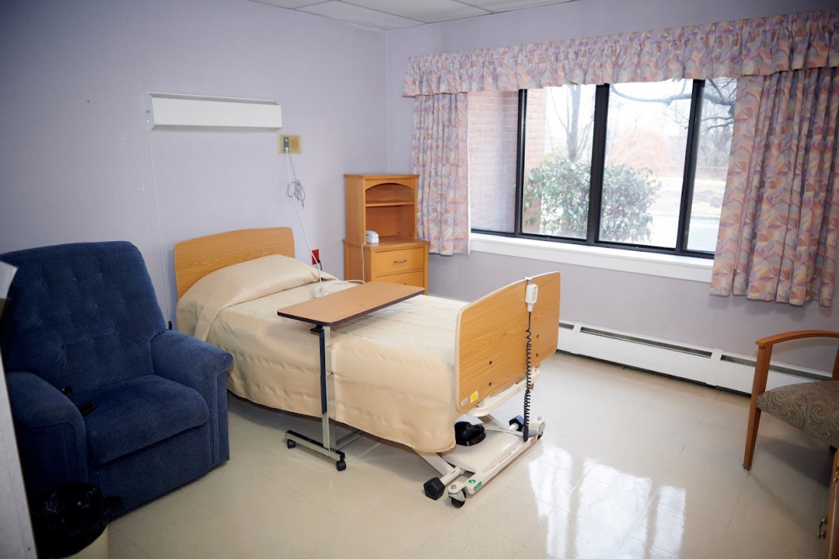 A clean and simple hospital room with a single bed, chair, and window providing natural light.