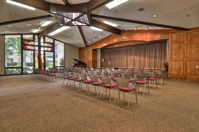 A modern conference room with a stage and rows of empty chairs, featuring a grand piano in the corner.