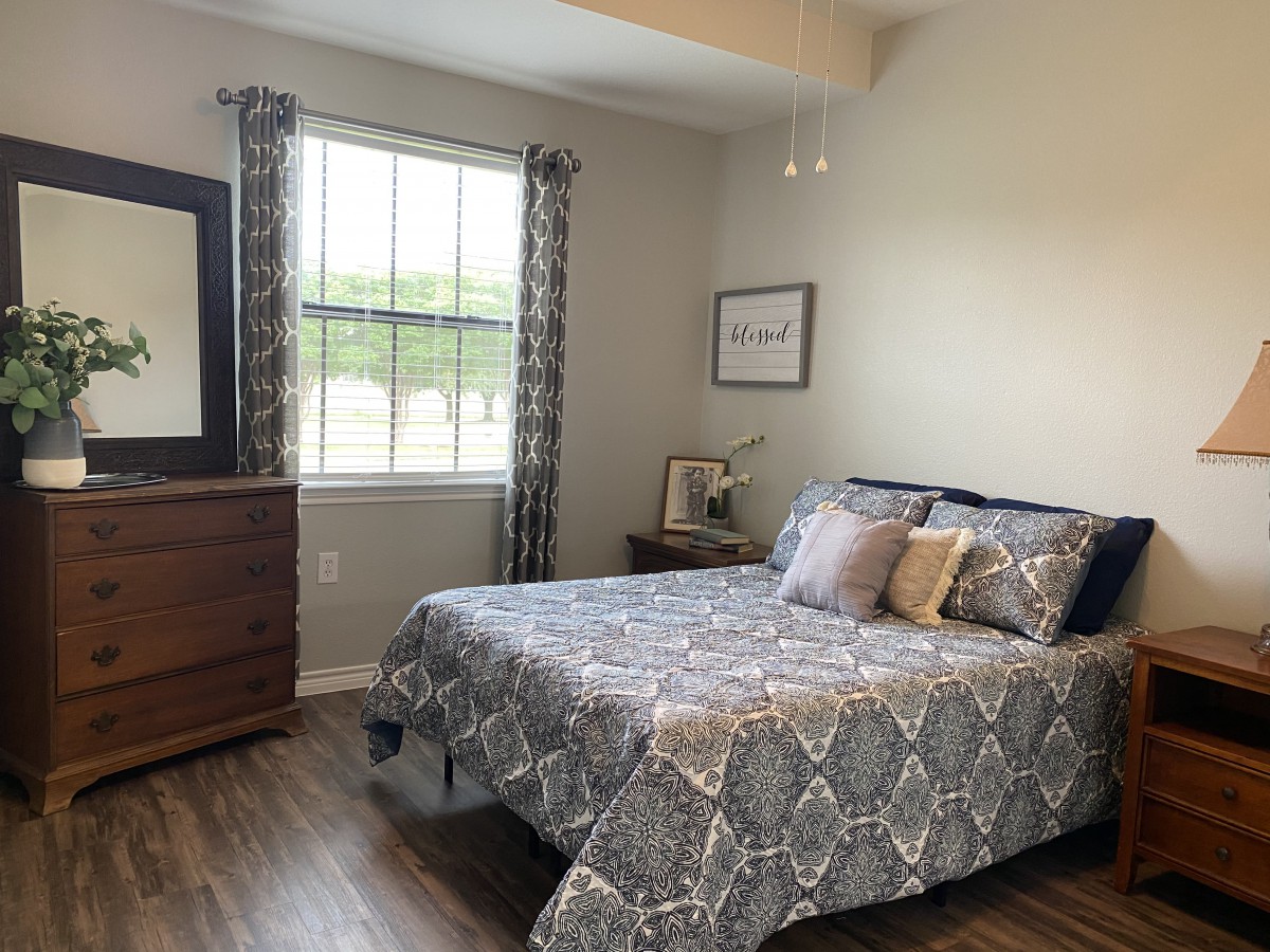 A cozy bedroom with a neatly made bed, wooden dresser, and window with patterned curtains.