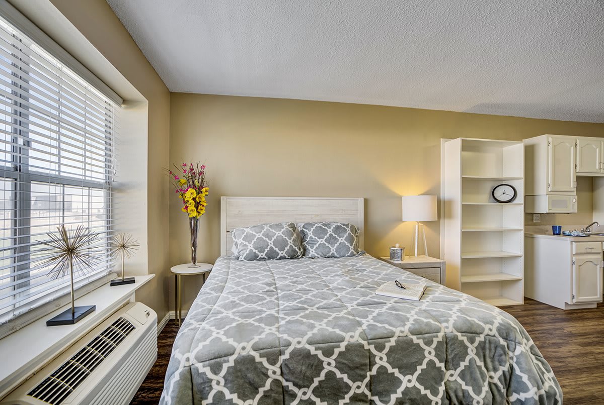 A cozy bedroom featuring a bed with patterned bedding, a lamp on a bedside table, a window providing natural light, and shelves for storage.