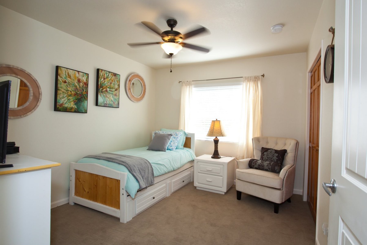 A cozy bedroom with a single bed, armchair, and bedside table under a ceiling fan.