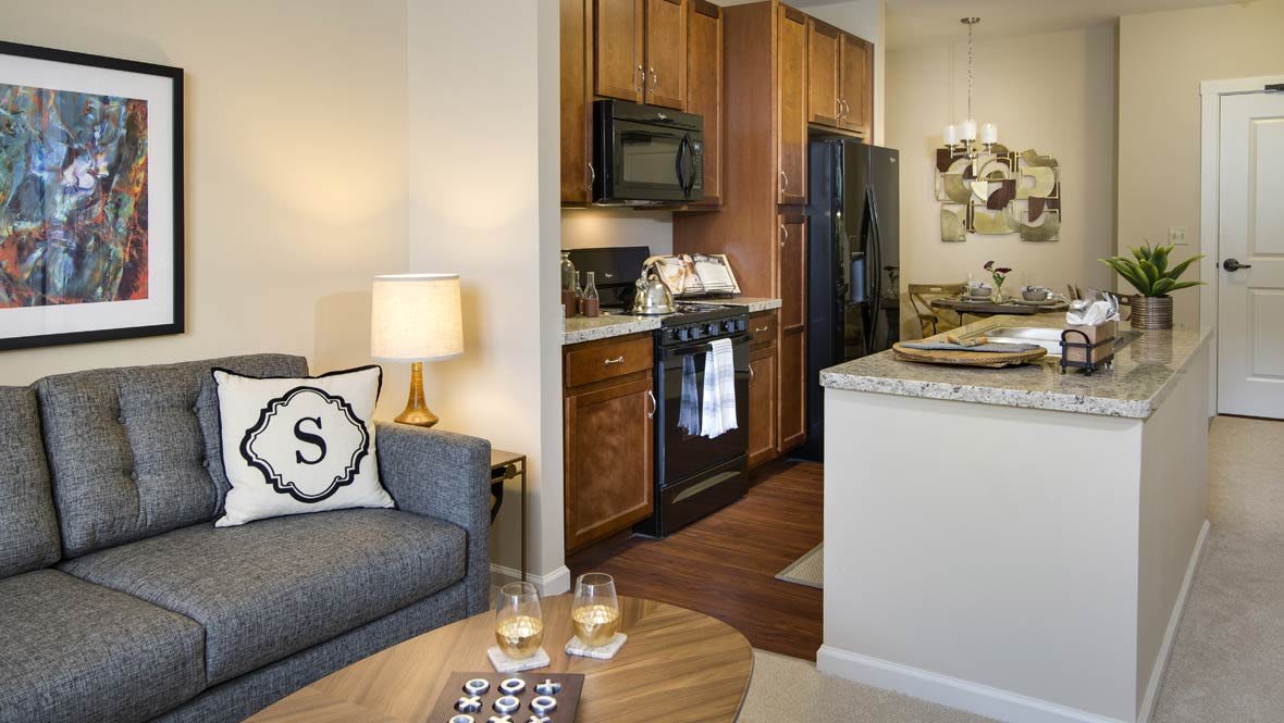 A cozy apartment interior featuring a living room with a sofa and lamp, adjacent to an open kitchen with wooden cabinets.